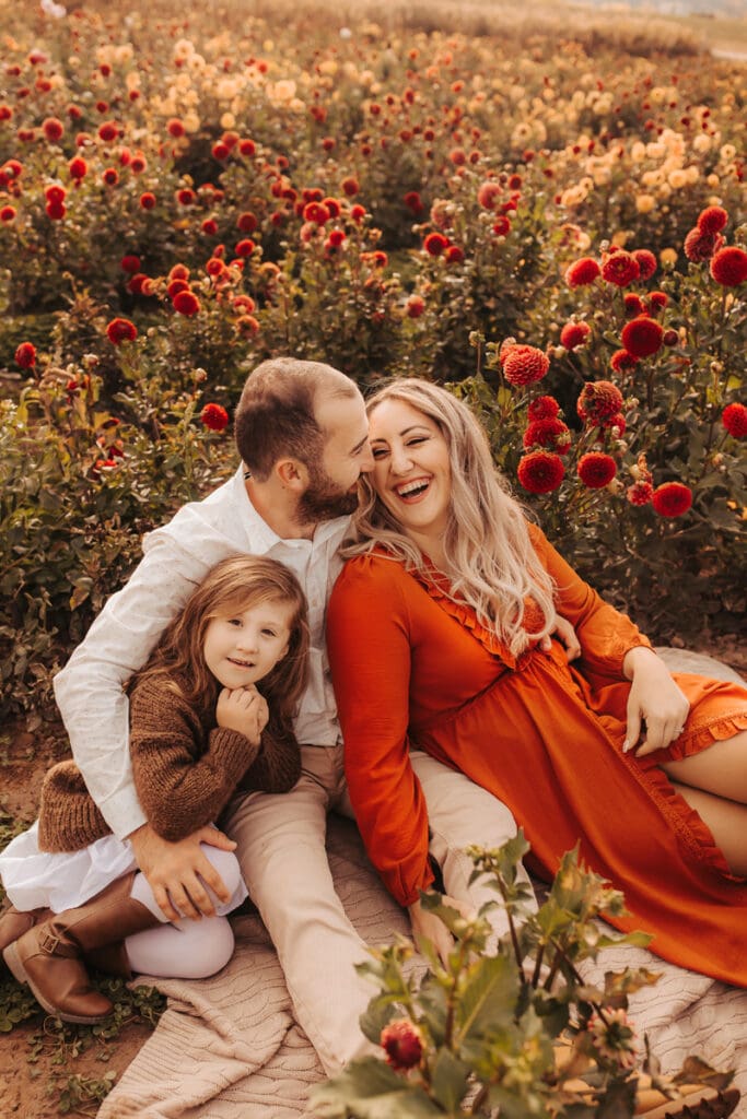 Family Photographer, a man kisses his wife as he holds their smiling daughter, they all sit in a field of flowers