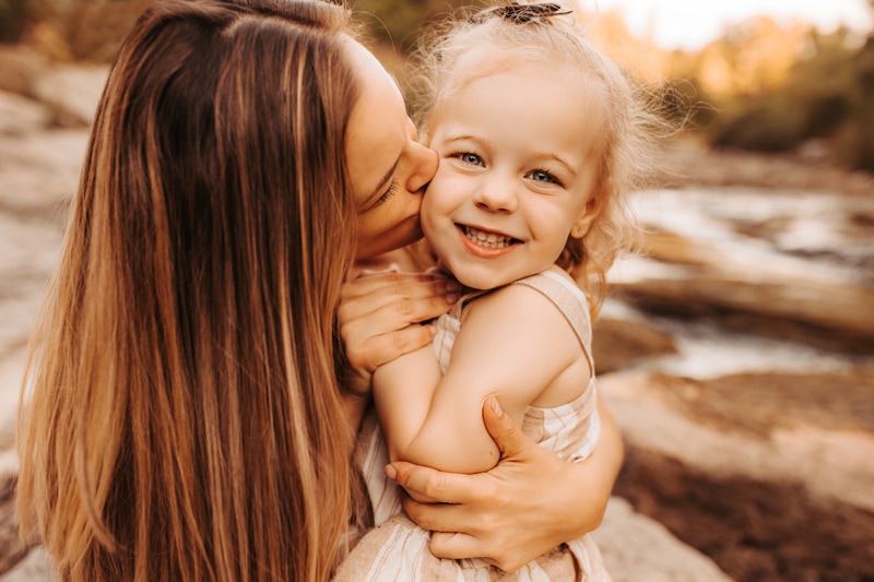 Family Photographer, a mother kisses her young daughter as she smiles near quiet streams