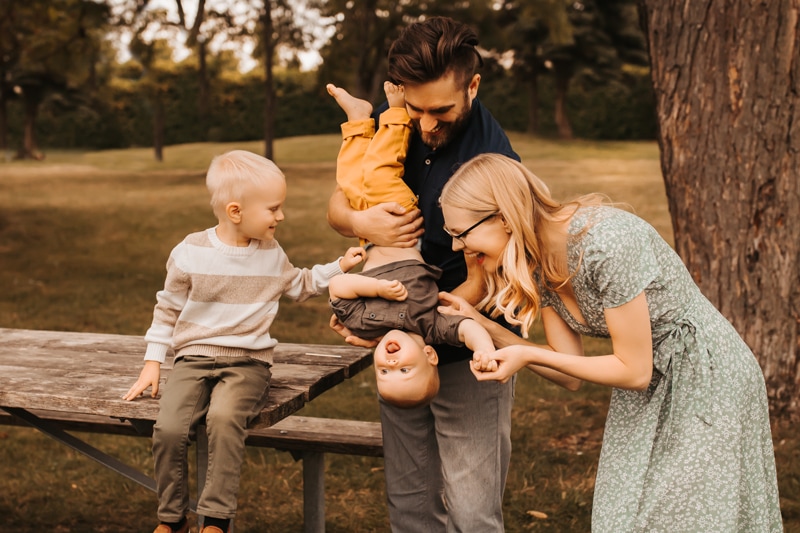 Family photographer, a father holds his son upside down playfully as mom and brother look on. They are outside at a park.