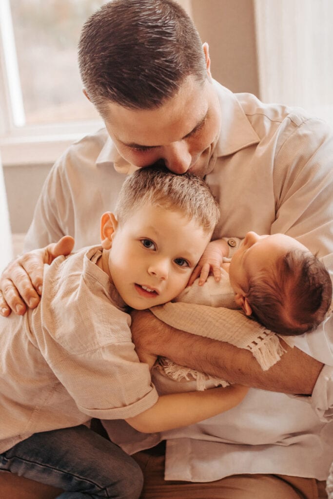 Newborn Photographer, a father holds his baby as the older brother embraces her also